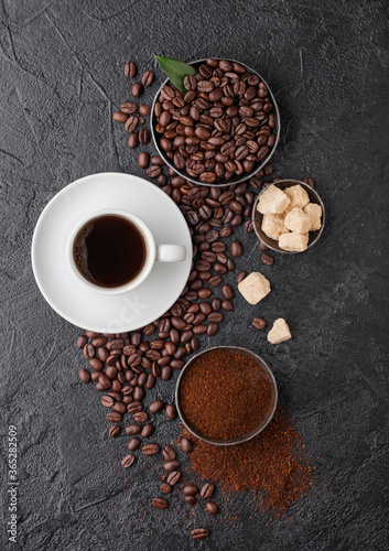 White cup of fresh raw organic coffee with beans and ground powder with cane sugar cubes with coffee tree leaf on black background. Top view