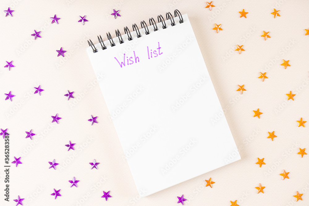 Spiral notebook lies on light background. On white sheet of paper, inscription in purple marker - Wish List. Around - purple and orange rhinestones in shape of stars. Top view, flat lay, copy space. Spiral notebook lies on light background. On white sheet of paper, inscription in purple marker - Wish List. Around - purple and orange rhinestones in shape of stars. Top view, flat lay, copy space.