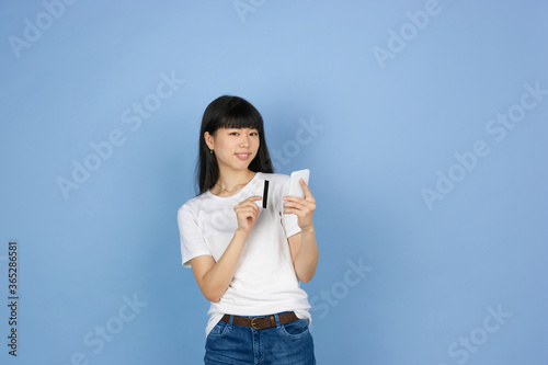 Showing phone and card, purchase. Portrait of young asian woman isolated on blue studio background. Beautiful cute girl in casual. Human emotions, facial expression, sales, ad, online shopping concept