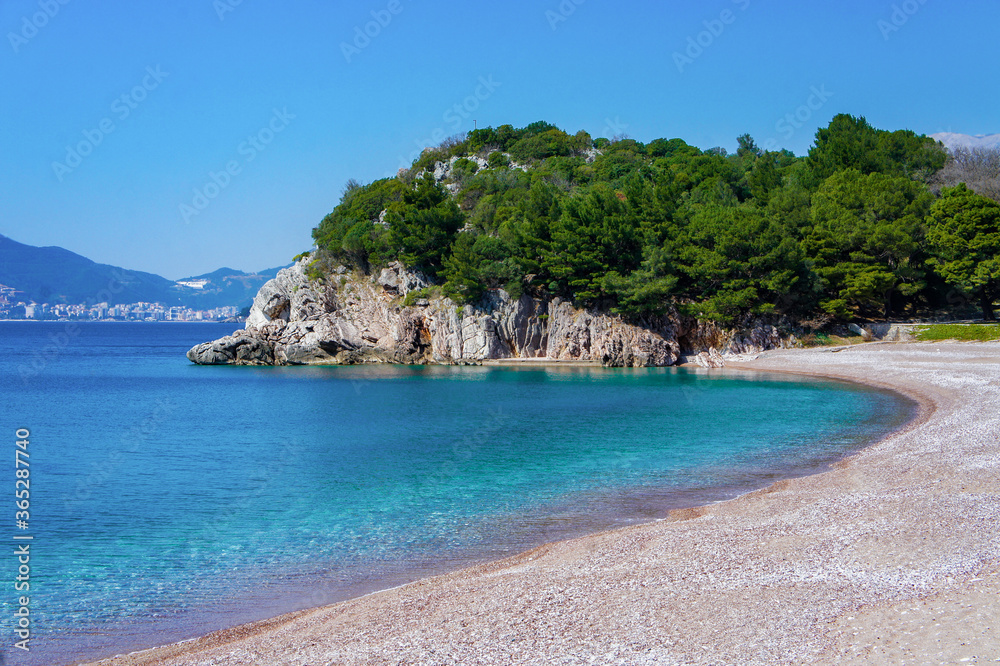 Mediterranean blue sea, beach with small orange pebbles, rocks with green pine trees.