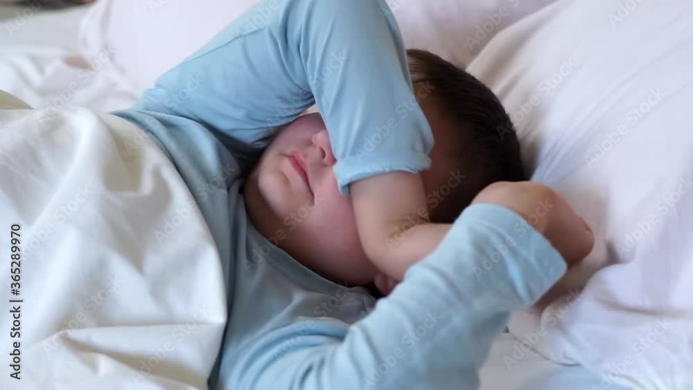 sleepy little child boy yawning and rubbing his eyes and stretching in white bed. happy kid wake up early in the morning under blanket before school. Bedtime, awakening toddler looking to camera. sleepy little child boy yawning and rubbing his eyes and stretching in white bed. happy kid wake up early in the morning under blanket before school. Bedtime, awakening toddler looking to camera.