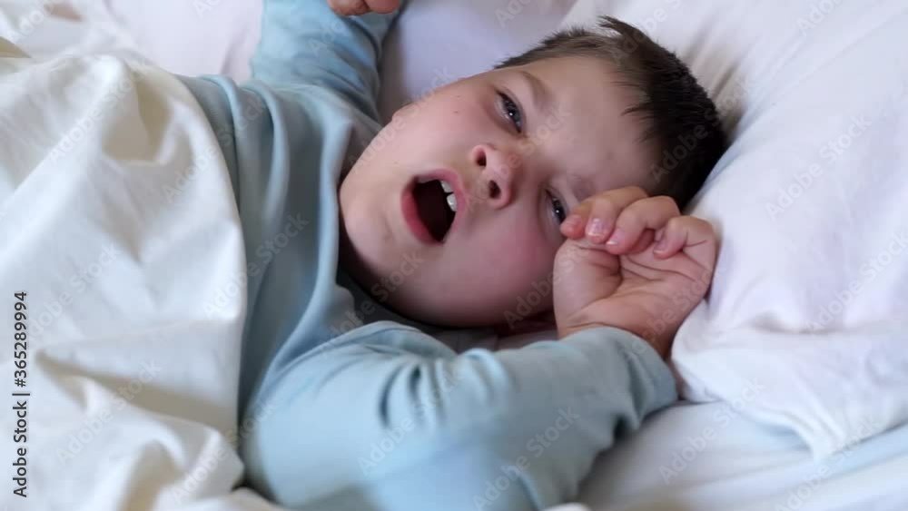 sleepy little child boy yawning rubbing eyes and stretching in white bed. happy kid wake up early in the morning under blanket and go to school. Bedtime, awakening toddler looking to camera.. sleepy little child boy yawning rubbing eyes and stretching in white bed. happy kid wake up early in the morning under blanket and go to school. Bedtime, awakening toddler looking to camera..