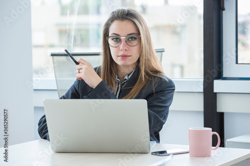 giovane manager bionda con occhiali da vista è seduta nella sua poltrona di lavoro davanti alla scrivania con il portatile e guarda con aria seria photo