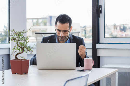 Manager uomo moro vestito con camicia blu e giacca nera porta degli occhiali da vista mentre lavora seduto nella sua postazione del suo ufficio 