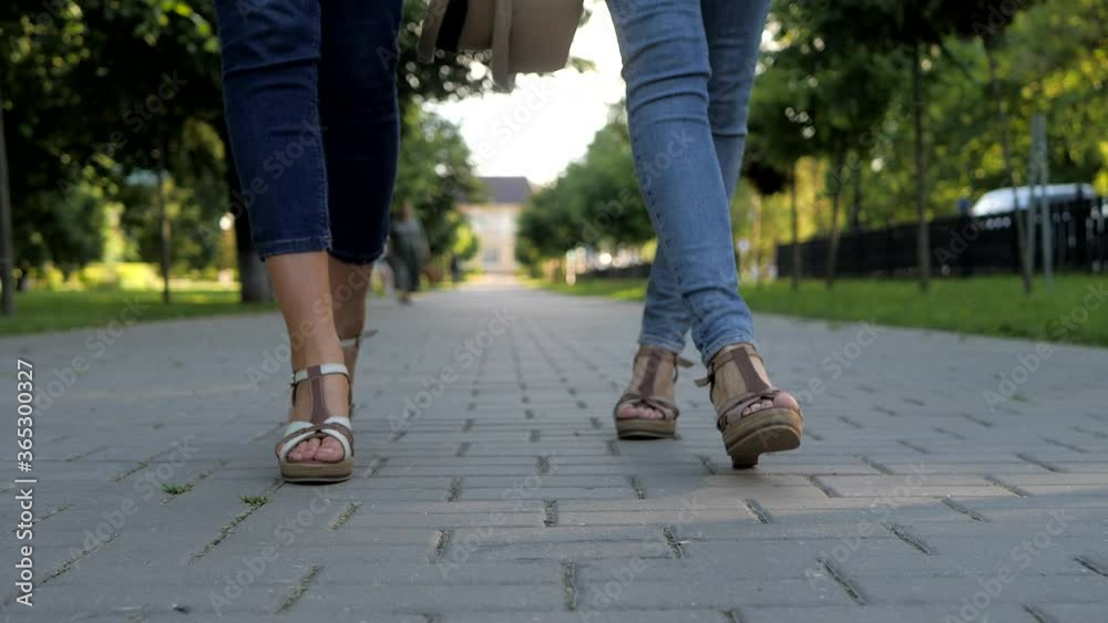 Two girlfriends walk together on a macadam in the Park. Feet shod in sandals close-up. Two girlfriends walk together on a macadam in the Park. Feet shod in sandals close-up.