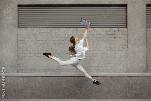 Paperwork. Jumping young woman in front of city building wall, on the run in jump high. Hurrying up to daily routine inspired and sportive. Young ballet dancer in casual clothes and sunshine. © master1305