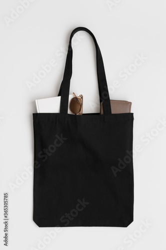 Black tote bag mockup with a book, wallet and glasses on a white table.
