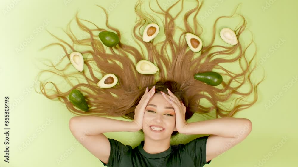 pretty surprised girl lying on green colored background with avocado fruits on long hair, young woman covers his mouth in delight, concept female health and beauty pretty surprised girl lying on green colored background with avocado fruits on long hair, young woman covers his mouth in delight, concept female health and beauty