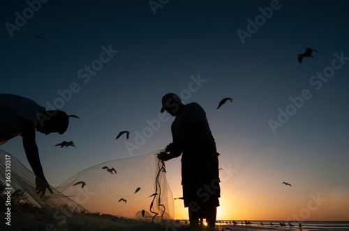 Fototapeta Two fishermen at sunset on the beach clean the fishing nets - in the background