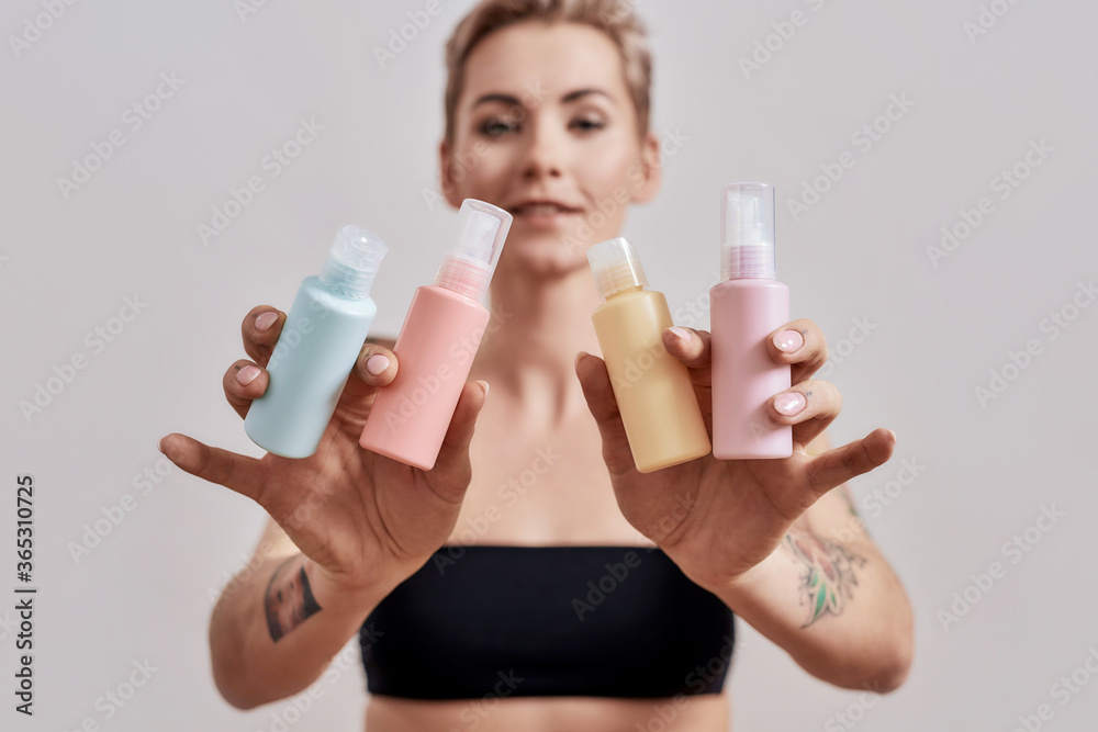 Portrait of beautiful tattooed woman with short hair holding plastic bottles with cosmetic, hair and skin care products, isolated over grey background Portrait of beautiful tattooed woman with short hair holding plastic bottles with cosmetic, hair and skin care products, isolated over grey background