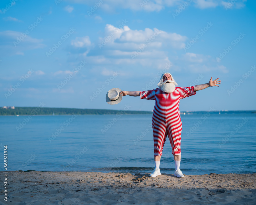 Elderly gray-haired man with a beard in a striped bathing suit and hat posing on the beach. Senior citizen on vacation by the lake.