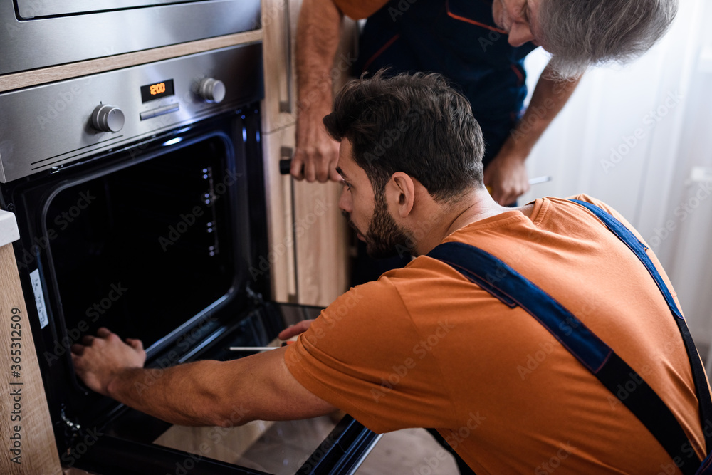 Obraz premium Close up shot of two repairmen, workers in uniform examining oven using screwdriver and flashlight indoors. Repair service concept