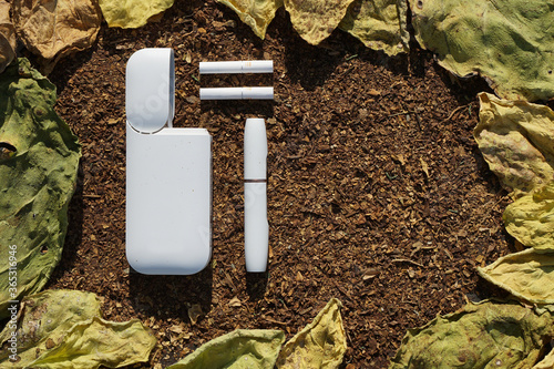White battery  with  individual stick e-cigarette and cigarette for it  lies on  dry  tobacco texture with green, yellow  leaves  of tobacco.