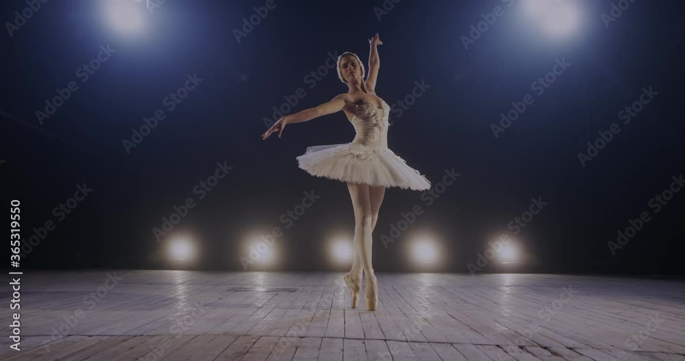 Graceful young female ballerina in tutu dancing charmingly and doing pas in light of blue light projectors on stage of theater. Camera zooming in woman dancer of ballet in lamps lights.