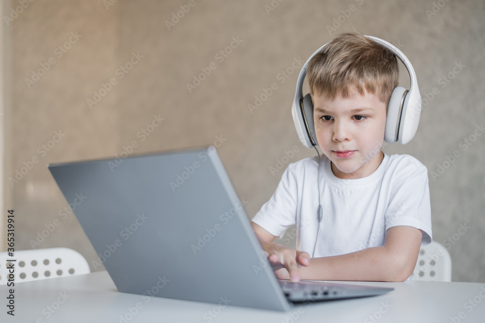 Cute little boy is watching cartoons or a movie on a laptop. Caucasian child sits at a table at home and listens to music on headphones. Cute little boy is watching cartoons or a movie on a laptop. Caucasian child sits at a table at home and listens to music on headphones.