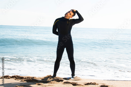 attractive and athlete  guy in wetsuit stretches on the beach before entering the sea with the surfboard