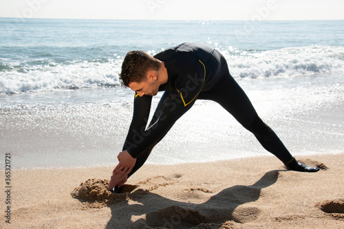 attractive and athlete  guy in wetsuit stretches on the beach before entering the sea with the surfboard