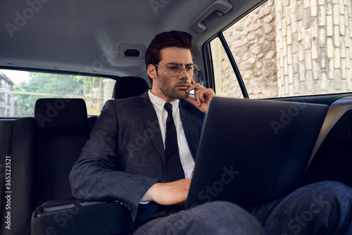 Confident young businessman seriously and thoughtfully looks at the laptop holding his chin with one hand while sitting in the car. © Roman