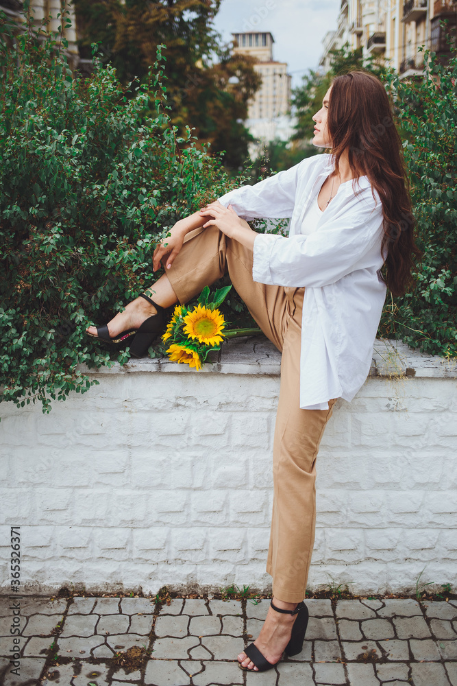 Young happy beautiful girl is walking in a shirt and pants with a bouquet of flowers of sunflowers on a city street