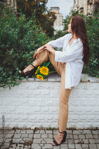 Obraz na płótnie Young happy beautiful girl is walking in a shirt and pants with a bouquet of flowers of sunflowers on a city street w salonie