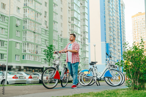Fototapeta Naklejka Na Ścianę i Meble -  Bearded man in casual clothes uses eco transport on the streets of the city, walks with a rented bicycle against the backdrop of a yard with apartment buildings. Tourist took a bicycle to the sharing