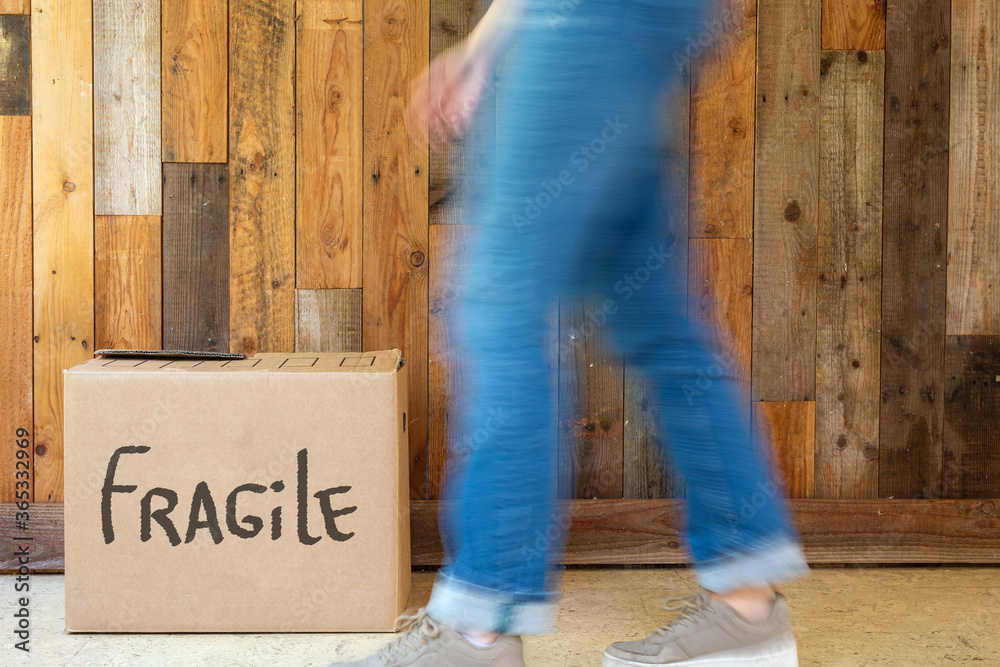 Young woman walks with moving box in new home near wooden wall, holding cardboard moving boxes on moving day retro design fragile written on box