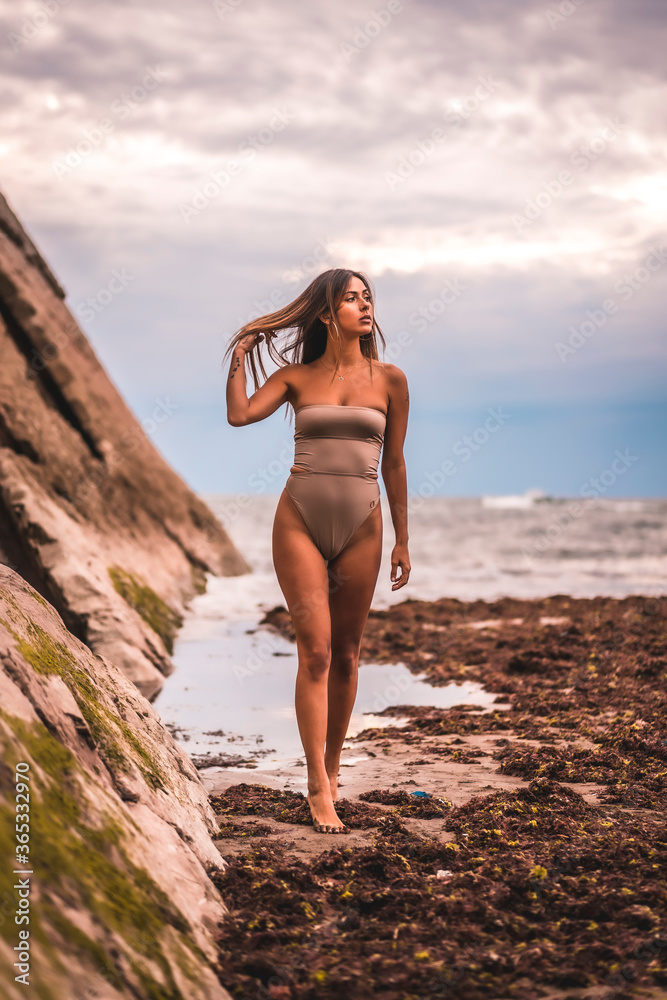 A dark-haired caucasian woman in a brown swimsuit on a natural background by the sea in the town of Zumaia, Gipuzkoa. Basque Country. In the summer sunset. Pretty young woman walking on the on coast