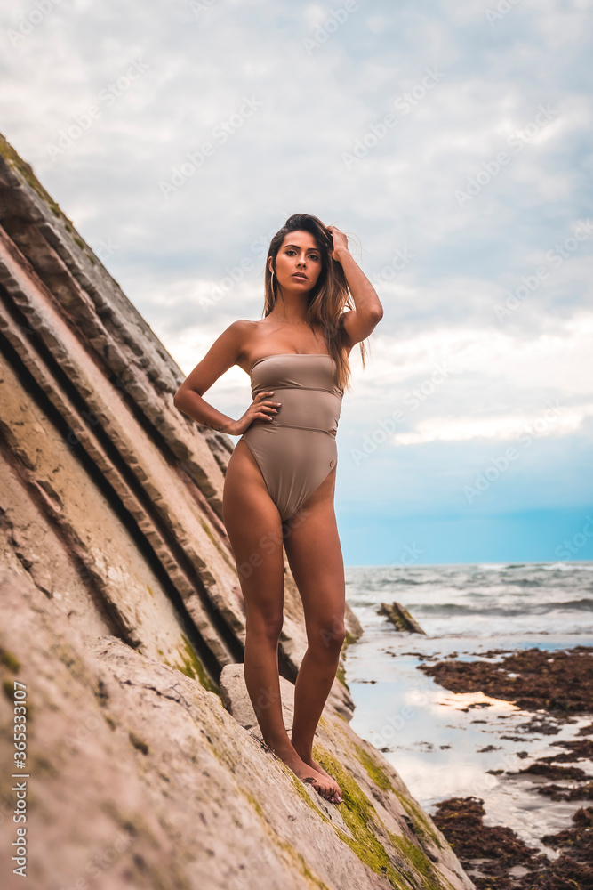 A dark-haired caucasian woman in a brown swimsuit on a natural background by the sea in the town of Zumaia, Gipuzkoa. Basque Country. In the summer sunset. On top of some rocks with the pensive A dark-haired caucasian woman in a brown swimsuit on a natural background by the sea in the town of Zumaia, Gipuzkoa. Basque Country. In the summer sunset. On top of some rocks with the pensive