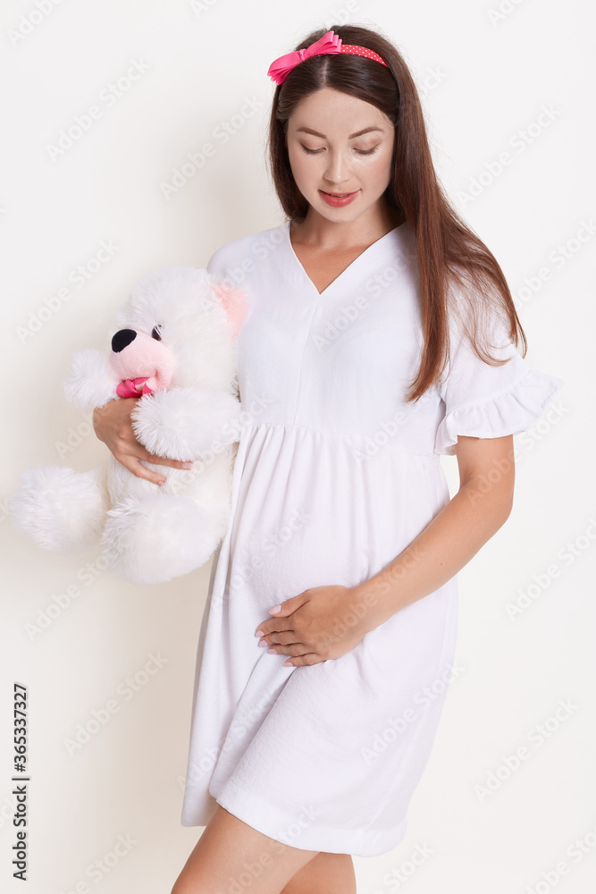 Lovely pregnant girl with teddy bear wearing white dress, dark haired female with soft bear, touching her belly and looking on it, looks happy and gently. Lovely pregnant girl with teddy bear wearing white dress, dark haired female with soft bear, touching her belly and looking on it, looks happy and gently.