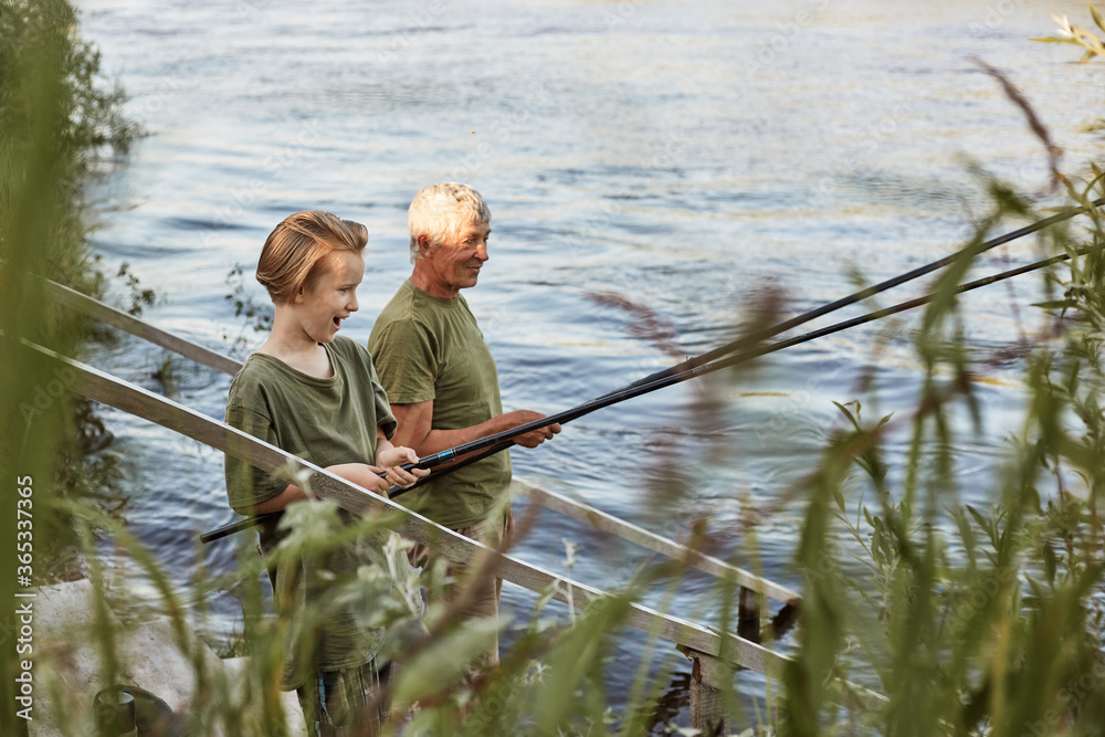 Fishing by lake, grandson and grandfather near river, holding fishing rods in hands, look excited and happy, stand laughing on wooden placing, family like fishing. Fishing by lake, grandson and grandfather near river, holding fishing rods in hands, look excited and happy, stand laughing on wooden placing, family like fishing.