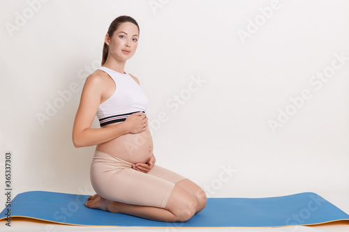 Pregnant woman doing exercise while sitting on blue yoga mat, touching her bare belly and looking at camera, expectant mother after pregnancy gymnastic.