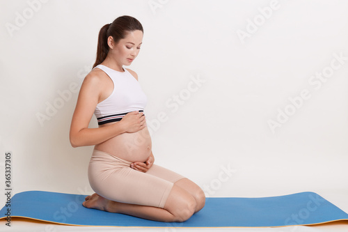 Relaxation for expectant mother and baby. Portrait of pregnant young woman touching her belly while sitting on blue yoga mat, female with ponytail wearing sporty top and leggins.