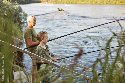 White haired pensioner with his grandson relaxing at blank of river or lake, senior man teaching boy to fish, family posing on wooden setting with fishing rods in hands.