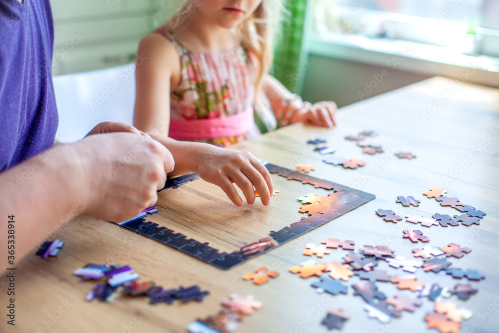 Fototapeta premium Father and daughter assemble the puzzle while sitting at the table. Games during rain and bad weather, quarantine. Family relaxing educational games at home.