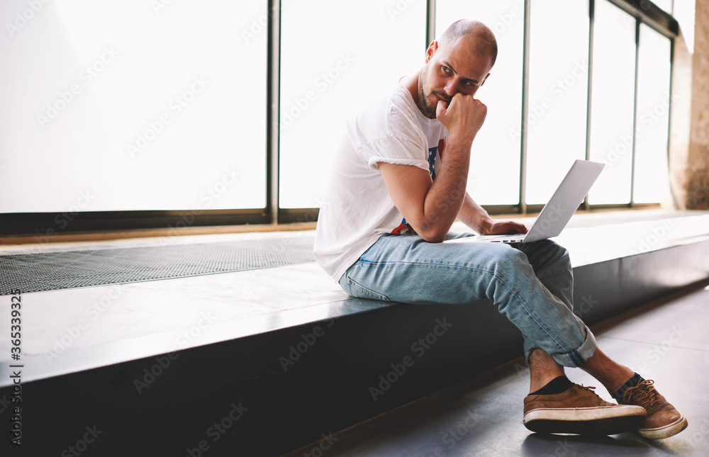 Young man looking thoughtful while working on laptop computer holding it on the knees, university student using notebook for write his coursework, freelancer sitting in modern interior thinking about