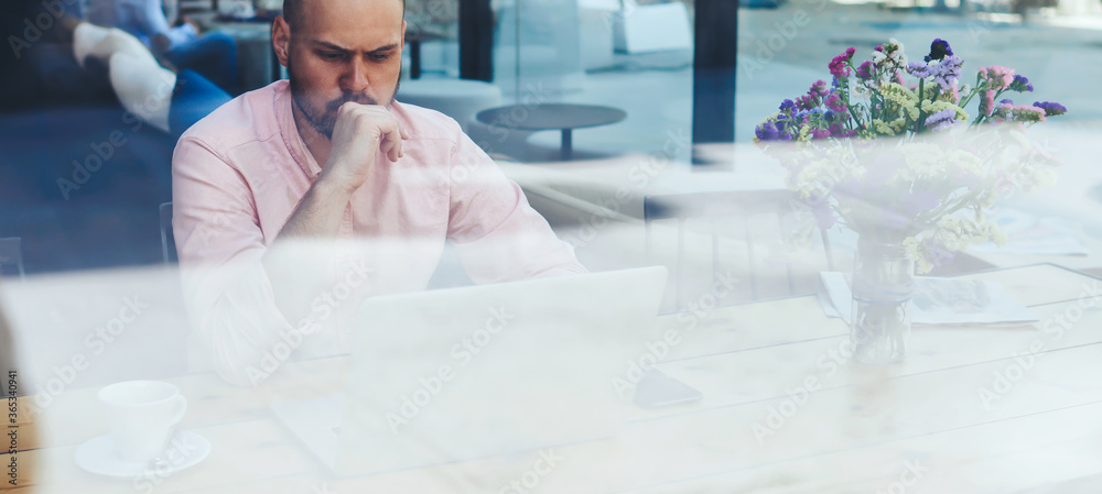 Fototapeta premium Puzzled and concerned male freelancer or student look to net-book, worried and thoughtful businessman or entrepreneur sitting front open laptop computer in modern coffee shop interior in urban setting