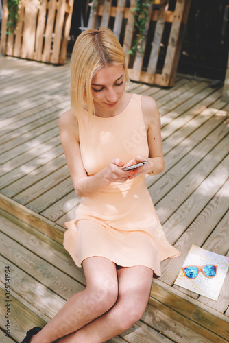 Lovely young woman sending text message with her mobile phone, charming hipster girl chatting on cell phone sitting outdoors in summer,female tourist connecting to wireless on telephone for navigation
