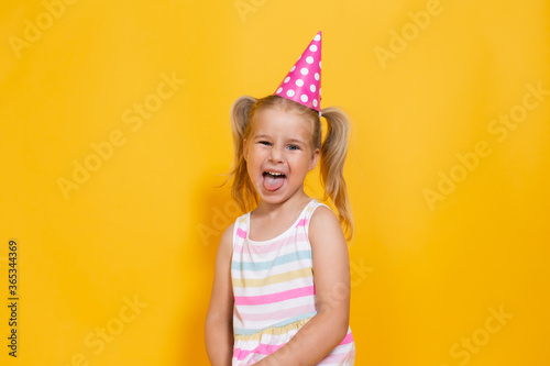 Happy birthday child girl with two ponytales in pink cap on colored yellow background shoing her tongue.