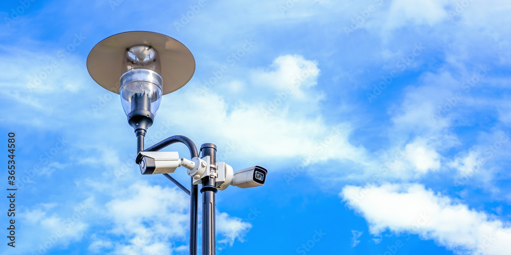 Two white surveillance cameras on the metal street light lamp post outdoor on blue sky background Two white surveillance cameras on the metal street light lamp post outdoor on blue sky background