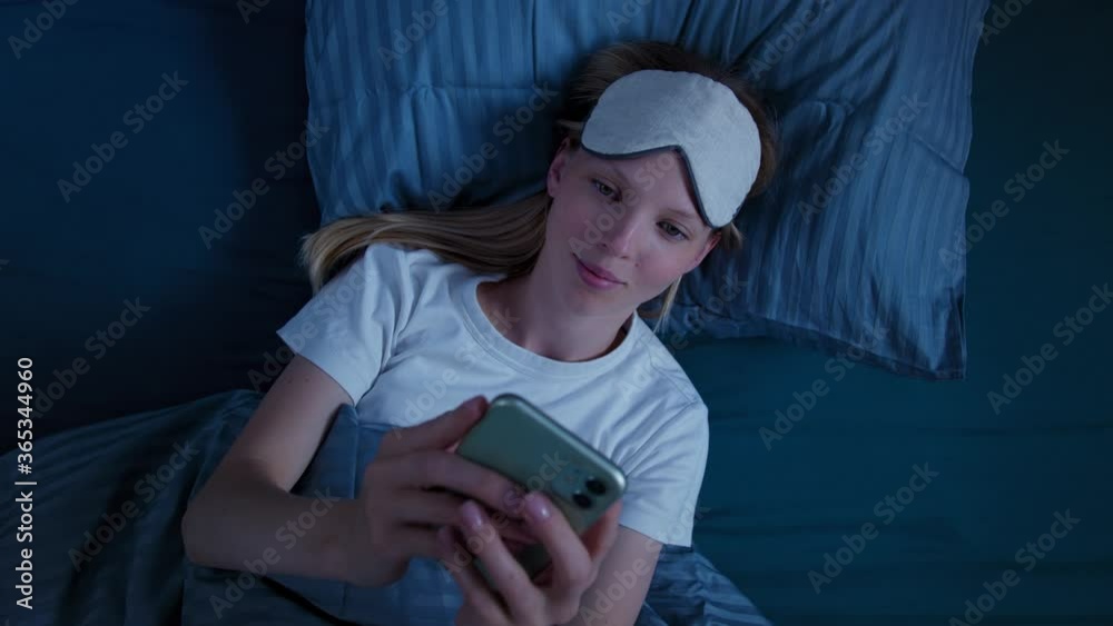 View top young beautiful woman lying in bed texting on phone social networks, reading news, getting ready for bed, wearing sleep mask View top young beautiful woman lying in bed texting on phone social networks, reading news, getting ready for bed, wearing sleep mask