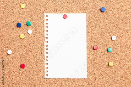 Flat lay of a blank paper with a red thumb tack holding it on a cork board with multi colored thumb tacks around