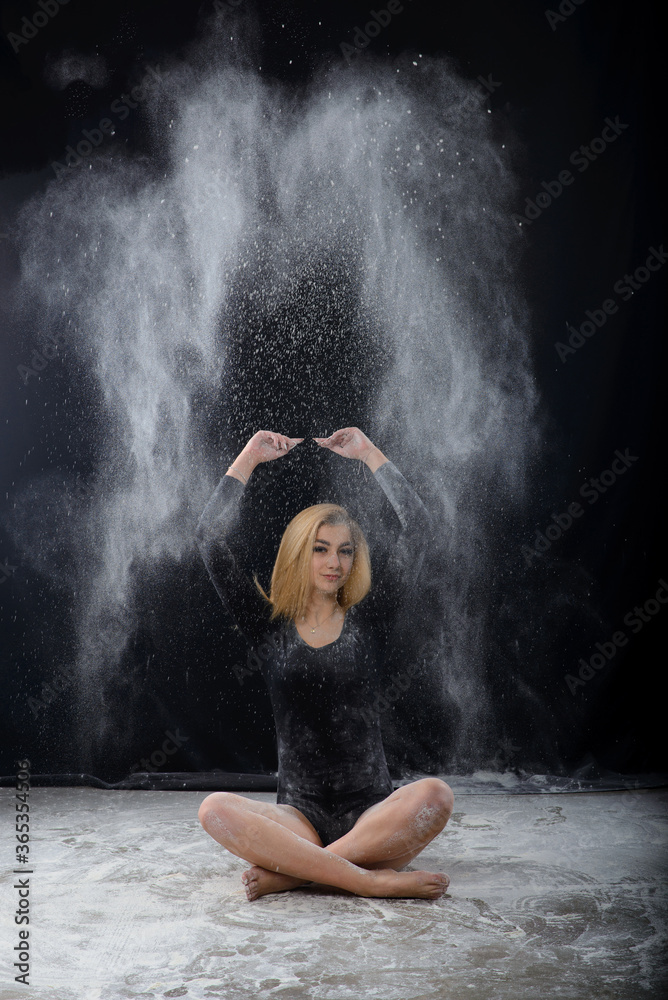 Fototapeta premium Piękna pulchna blondynka ubrana w czarne gimnastyczne body pokryte chmurami lecącego białego proszku skacze tańcząc siedząc w ciemności. Artystyczne zdjęcie koncepcyjne i reklamowe.