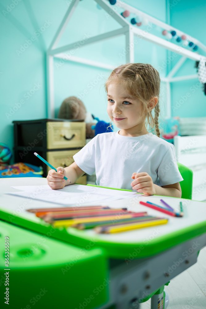 A girl in a white T-shirt sits in her room at the table and draws with colored pencils A girl in a white T-shirt sits in her room at the table and draws with colored pencils