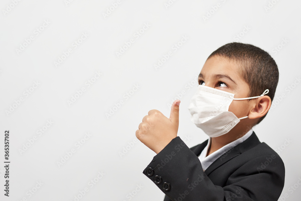 Happy schoolboy in face mask. Mixed-race kid shows thumbs-up gesture between pile of books. Back to school after covid-19. School year start after long delay due to coronavirus Happy schoolboy in face mask. Mixed-race kid shows thumbs-up gesture between pile of books. Back to school after covid-19. School year start after long delay due to coronavirus
