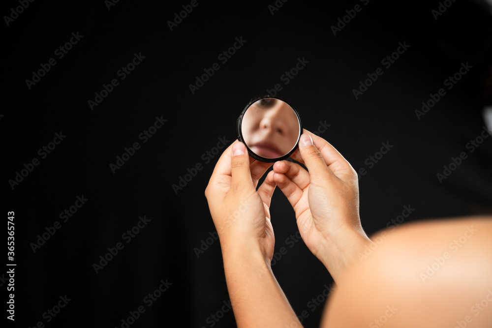 Fototapeta premium Hispanic woman looking at herself in a mirror with black background - close up hands holding a small mirror with the reflection of a face in it - reflection of her face