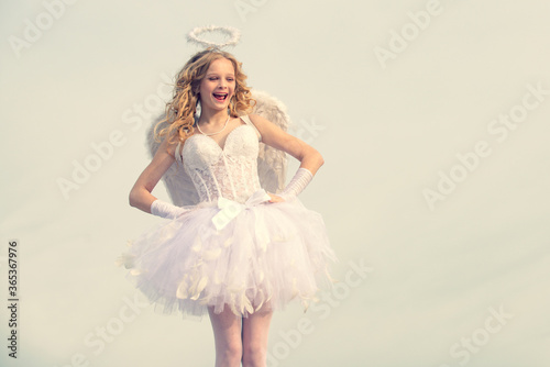 Innocent Girl with angel wings standing with bow and arrow against blue sky and white clouds. Portrait of a cupid little girl pray. Child with angelic character.