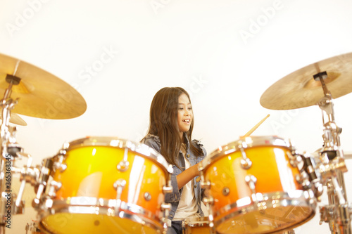 Fototapet A cute Asian elementary school girl with long hair and a denim jacket is in a good mood when learning to play a drum in a classroom with a white wall
