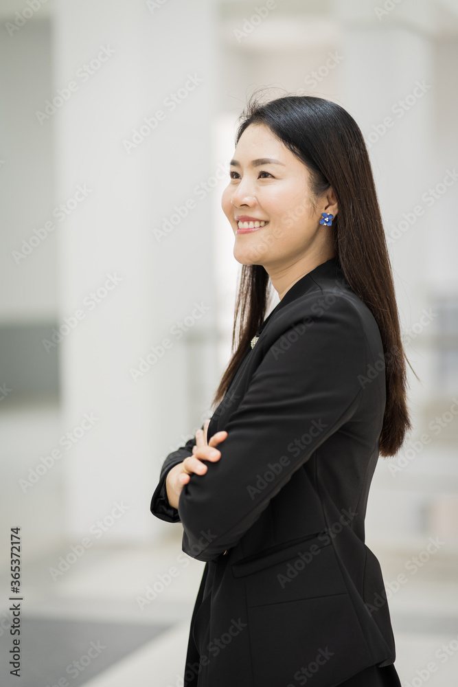 Portrait of a cheerful middle-age businesswoman in business suit stands in the company building with confidence arms crossed. Modern business woman in the office with copy space. Business stock photo. Portrait of a cheerful middle-age businesswoman in business suit stands in the company building with confidence arms crossed. Modern business woman in the office with copy space. Business stock photo.