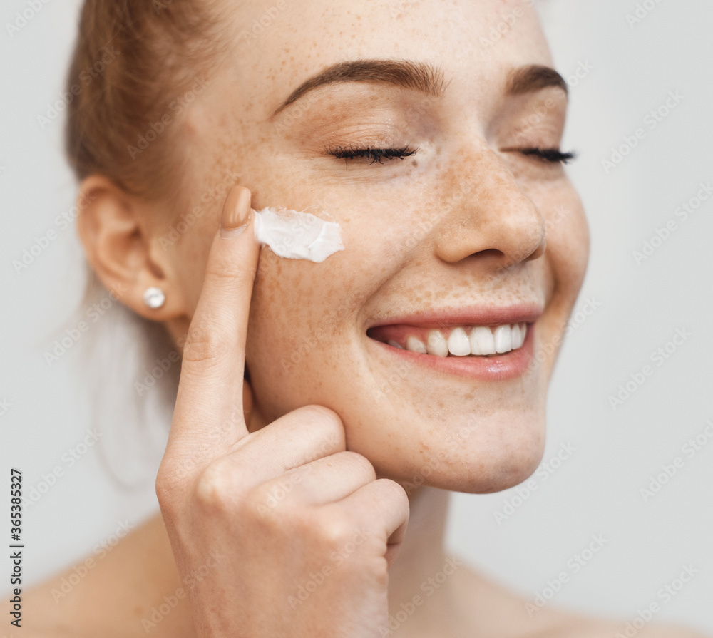 Freckled caucasian ginger lady applying on face anti aging cream and smile on a white studio wall - obrazy, fototapety, plakaty 