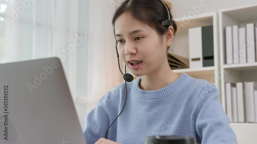 Smiling Asian business woman in casual clothes and wearing microphone headset of customer support phone operator at workplace.	